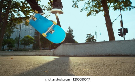 LOW ANGLE, CLOSE UP: Unrecognizable male skater does a kickflip in the sunlit urban park. Cool shot of blue skateboard flipping and turning under unknown man's feet. Man skating in the conrete park.