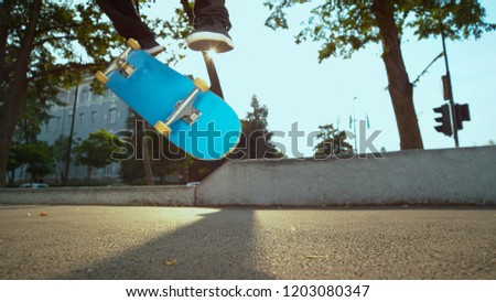 LOW ANGLE, CLOSE UP: Unrecognizable male skater does a kickflip in the sunlit urban park. Cool shot of blue skateboard flipping and turning under unknown man's feet. Man skating in the conrete park.