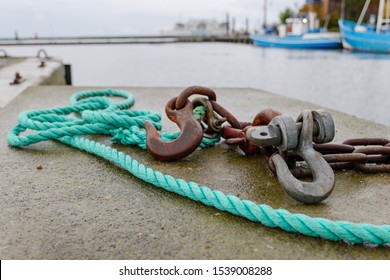 Low angle and close up view of rusty hook and green nylon rope on concrete floor of waterside at harbour   in Denmark with blur background of boats and cloudy sky.
