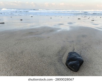 Low angle close-up of dark pebbles resting on smooth wet sand with ocean waves blurred in the background