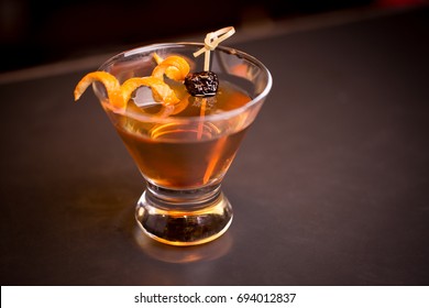 Low Angle Closeup Of Gourmet Old Fashion Martini Drink In Stemless Martini Cocktail Glass Filled With Bourbon And Brandy Garnished By Delicately Spiral Orange Peel And Preserved Cherry Countertop Bar