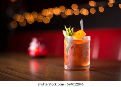 Low Angle Closeup Of Gourmet Old Fashion Craft Cocktail Of Sweet Bourbon Whiskey Drink Garnished By Orange Peel And Rosemary On Table Of Red Leather Diner Booth With Blurry Bokeh Restaurant Background