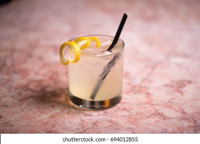 Low Angle Closeup Of Gourmet Old Fashion Mixed Drink Craft Cocktail Of Sweet Lemonade Vodka And Gin Martini Garnished By Delicately Spiraled Lemon Peel With Straw On Pink Marble Bar Countertop Bar