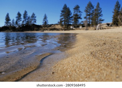  Low angle close-up of a pristine sandy beach shoreline on a calm lake with evergreen pine trees under a clear blue sky.