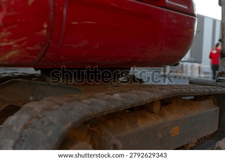 Low angle close-up of a red tracked construction machine showing the metal undercarriage, worn track parts, and an industrial worksite in the background