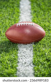 A low angle close-up view of a leather American Football sitting in the grass on a white yard line.