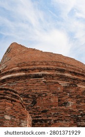 Low angle close-up view of the weathered red brick base of a ruined ancient stupa at Wat Kudi Dao in Ayutthaya, Thailand. The historic architecture stands against a bright blue sky with wispy clouds