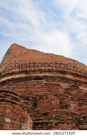 Low angle close-up view of the weathered red brick base of a ruined ancient stupa at Wat Kudi Dao in Ayutthaya, Thailand. The historic architecture stands against a bright blue sky with wispy clouds