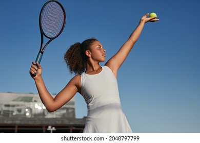 Low Angle Of Concentrated Sporty Teen Female With Curly Hair Preparing To Hit Ball, With Racket While Playing Tennis Game On Court Against Blue Sky