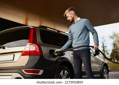 Low angle of concentrated young bearded male driver in smart casual clothes filling fuel into modern SUV car with gas pump nozzle at station