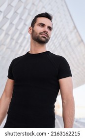 Low Angle Of Confident Young Bearded Arab Guy With Dark Hair In Black T Shirt, Smiling And Looking Away While Standing On Street Near Modern Geometric Building In Daytime