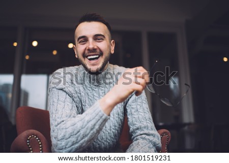 Low angle of content bearded man in casual wear smiling and looking at camera while enjoying time in cozy cafe