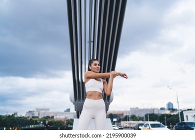 Low Angle Of Content Young Ethnic Female Athlete With Long Dark Hair In Sportswear And True Wireless Earphones Stretching Arms And Looking Away During Workout On Modern City Street