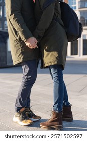 Low angle cropped view of a young adult couple wearing jeans and parka coats standing on a paved street holding hands