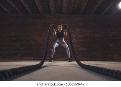 Low angle distant view of caucasian fit woman dressed in sportsoutfit exercising with battle ropes at gym.