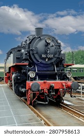 A low angle dramatic frontal view of a classic black steam locomotive with prominent red front beam and headlights standing on a platform track system.