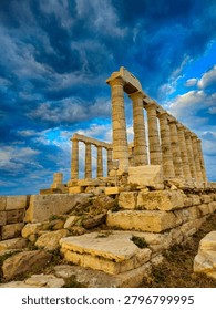 Low angle dramatic view of the ancient stone foundations and pillars of the Poseidon Temple at Cape Sounion under stormy sunset clouds.