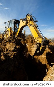 Low angle dramatic view of excavator bucket digging deep trench in soil. Heavy construction equipment shaping ground for foundation works.