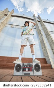 Low angle fashion shot of young Asian man standing on boombox in urban city setting against sky