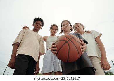 Low angle of four serious intercultural boys and girls in activewear looking at camera while one of them holding ball for playing basketball - Powered by Shutterstock