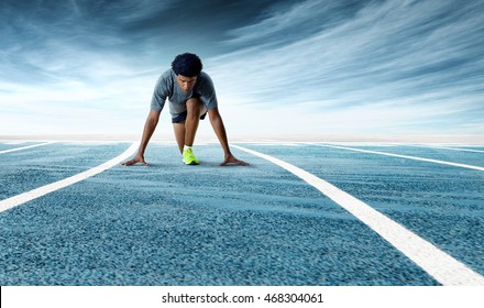 A low angle front shot of a determined African American sprinter preparing to start racing on a blue empty running track against dramatic sky background