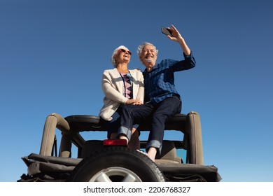 Low angle front view of a senior Caucasian couple sitting on the roof of their car at the beach in the sun, embracing, smiling and using a smartphone to take a selfie - Powered by Shutterstock