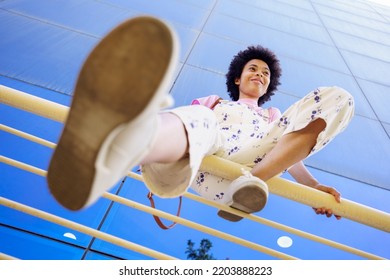 Low angle full body of glad African American female in casual clothes and sneakers looking away while sitting on railing near modern building - Shutterstock ID 2203888223