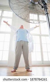 Low Angle Full Length Shot Of A Senior Man With Arms Raised By The Fan Indoors Seeking Respite From The Heat.