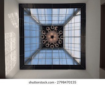 Low angle full shot of a square glass skylight ceiling with a large vintage chandelier hanging in the center, showcasing modern architecture design.
