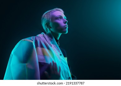 Low angle of futuristic young woman in translucent raincoat with short hair looking at colorful neon light against dark background - Powered by Shutterstock