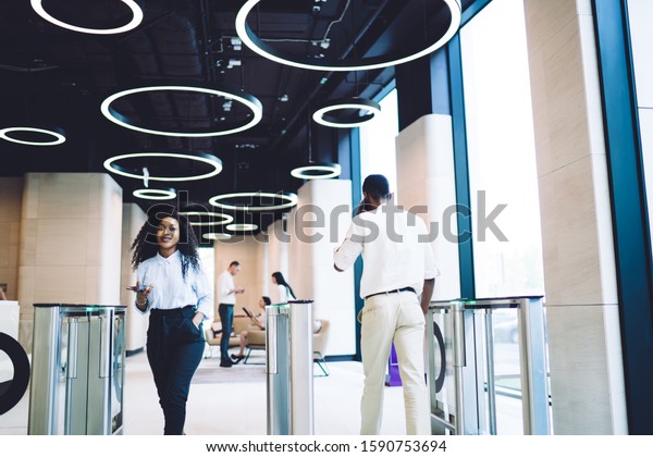 Low angle of glad African American staff\
in stylish clothes passing through turnstile while using smartphone\
in hall of contemporary business\
center