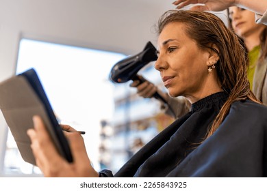 Low angle of hairdresser drying hair of female client using modern tablet while working as designer remotely during beauty routine - Powered by Shutterstock