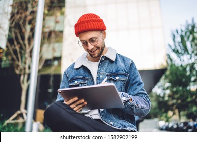 Low angle of handsome man in glasses sitting on street near glass skyscraper while working on tablet and doing graphic design - Powered by Shutterstock