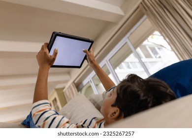 Low angle of happy biracial boy lying on couch using tablet with copy space on screen in living room. Childhood, free time, communication, entertainment, lifestyle and domestic life unaltered. - Powered by Shutterstock