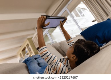 Low angle of happy biracial boy lying on couch using tablet with copy space on screen in living room. Childhood, free time, communication, entertainment, lifestyle and domestic life unaltered. - Powered by Shutterstock