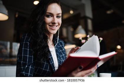 Low Angle Of Happy Female In Trendy Wear Smiling And Looking At Camera While Sitting In Cafe And Reading Book