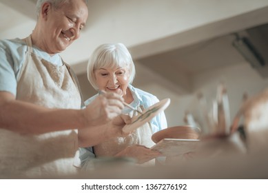 Low angle of happy male artist in apron painting decorative plate with paintbrush while making ornament on earthenware in workshop. Gray-haired wife is looking at his work indoors. Ceramic art concept - Powered by Shutterstock