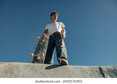 Low angle of happy male skateboarder holding skateboard standing at skate park in sunny day - Powered by Shutterstock