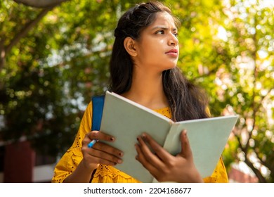 Low Angle Image Of Beautiful Indian Female College Student Holding A Book And Thinking While Looking Away At Day Time In Outdoor.   