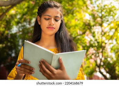 Low Angle Image Of Beautiful Indian Female College Student Reading A Book At Day Time In Outdoor.   