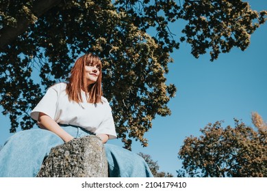 Low Angle Image Of A Body Positive Modern Girl On Jeans And White Shirt With Copy Spac, Trees And Sky Background. Modern Fashion Campaign Ad Concept. Bright Girls World Feminism And Strong Woman