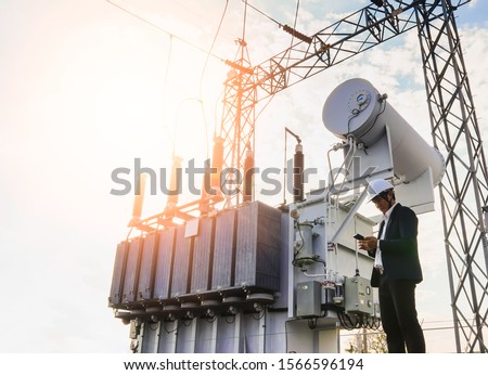 A low angle image of a businessman wearing a black suit, standing looking at a large power transformer with blue sky to be background, Concept about business people who want to invest in energy.