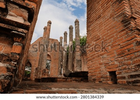 Low angle interior view of ancient red brick walls and weathered pillars inside the ruined assembly hall at Wat Kudi Dao in Ayutthaya, Thailand, looking up towards a cloudy blue sky and green trees