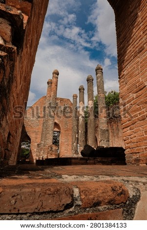 Low angle interior view of the historic brick assembly hall ruins at Wat Kudi Dao in Ayutthaya, Thailand. Weathered brick columns and walls stand tall against a cloudy blue summer sky with green trees visible through the arches