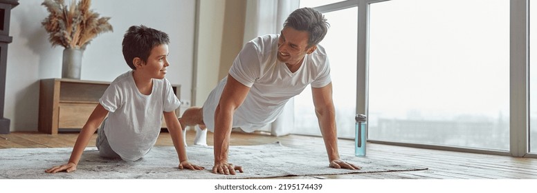 Low angle of jolly son enjoying workout with dad and doing plank together near window - Powered by Shutterstock