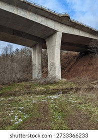 A low angle of a large concrete bridge going over a ravine with snow patches on the ground in the forest below.