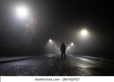 Low Angle, Looking Up At A Hooded Figure Standing In The Middle Of The Road On An Atmospheric Foggy Winters Night. UK.