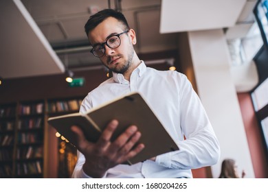 Low Angle Of Male In White Shirt And Spectacles Reading Book And Frowning While Standing On Blurred Background Of Cozy Bookshop