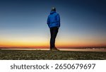 From low angle, man in a blue jacket stands alone on pier, looking out at distant city lights across the water during the tranquil blue hour after sunset, evoking a sense of calm and urban solitude.