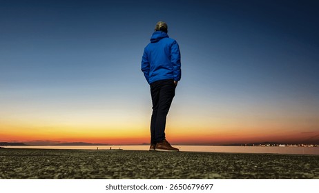 From low angle, man in a blue jacket stands alone on pier, looking out at distant city lights across the water during the tranquil blue hour after sunset, evoking a sense of calm and urban solitude.